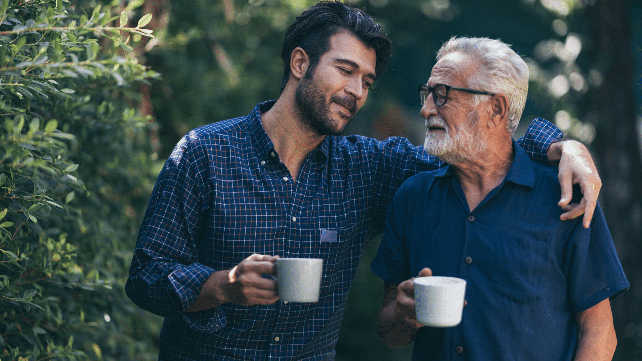 A younger man and an older man stand outdoors in a leafy garden, smiling at each other with their arms around one another’s shoulders, each holding a white mug.